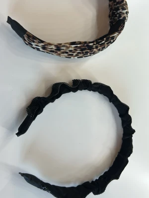 Två snygga diadem, svart & mönstrat - Säljer diadem! 10kr/styck! ett svart med glittrande detaljer och rynkad design, samt ett mönstrat i beige, svart och brunt med mjuk textil. Perfekta accessoarer för att lyfta din look och hålla håret på plats.