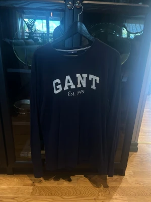 Mörkblå långärmad tröja från GANT - Snygg mörkblå långärmad tröja från GANT med vit logga och texten 'Est. 1949' på bröstet. Tröjan har rund halsringning och är tillverkad i mjukt material som passar perfekt till en avslappnad stil. Går att pruta.