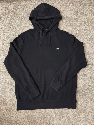 Svart hoodie från Lee - Svart hoodie från Lee med minimalistisk design och liten vit logga på bröstet. Klassisk modell med huva och dragsnören samt ribbade muddar vid ärmslut och nederkant. Perfekt för en avslappnad och stilren look.