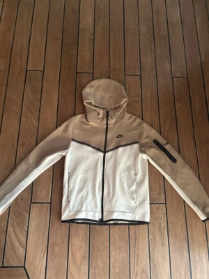 Nike tech - Snygg hoodie från Nike i beige och vitt med svart dragkedja och detaljer. Hoodien har huva, två sidofickor med dragkedja och en extra ficka på ena ärmen. Tillverkad i mjukt material som passar perfekt för en avslappnad stil.