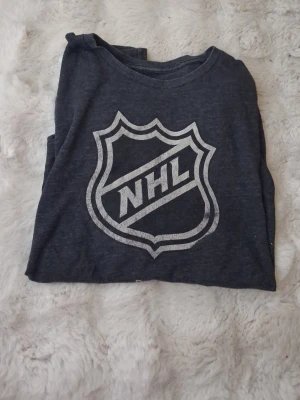 Mörkgrå NHL t-shirt med tryck - Snygg mörkgrå t-shirt med stort NHL-tryck på bröstet. Klassisk rund halsringning och korta ärmar. Perfekt för dig som gillar hockey och vill ha en sportig stil. Tillverkad i mjukt bomullsmaterial för skön känsla.