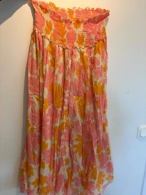 Blommig långklänning från Poppyfield - Långklänning från Poppyfield med blommigt mönster i rosa, orange och vitt. Klänningen har smock upptill och ett flowy fall, perfekt för varma dagar. Tillverkad i ett lätt och luftigt material som känns skönt mot huden.