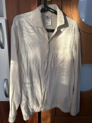 Beige långärmad skjorta från H&M - Säljer en stilren beige skjorta från H&M i mjukt material. Skjortan har klassisk krage, bröstficka och knäppning framtill. Perfekt för dig som gillar en enkel och clean look. Långärmad modell som passar året runt.