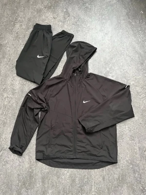 Äkta Svart Nike milner träningsset med huva - Säljer ett svart träningsset från Nike med jacka och byxor. Jackan har huva, dragkedja och Nike-logga på bröstet. Byxorna har resår i midjan och vid benslut samt vit Nike-logga på låret. Perfekt för träning eller chill. Materialet är lätt och andas. Säljer denna för att jag behöver rensa garderoben.