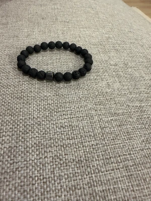 Svart armband med lavapärlor - Snyggt armband med svarta lavapärlor och en metallisk detalj. Pärlorna är runda och har en matt, lite porös yta som ger en cool och unik look. Perfekt accessoar för dig som gillar stilrena och trendiga smycken.