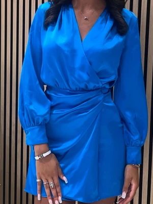 Blå omlottklänning i satin - Snygg blå omlottklänning från Zara i glansigt satinmaterial. Klänningen har lång ärm med lätt puff och markerad midja med drapering. Perfekt för dig som vill sticka ut med en färgstark look. Den har en dragkedja vid sidan av klänningen.