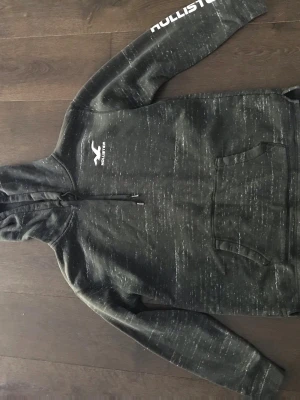 Svart hoodie från Hollister - Säljer en svart hoodie från Hollister med vit logga på bröstet och text på ärmen. Hoodien har huva med dragsko och en stor magficka. Materialet är mjukt och bekvämt, perfekt för chill dagar. Snyggt melerad design som ger en avslappnad vibe.