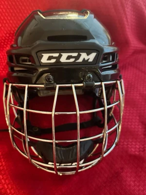 Svart hockeyhjälm med galler CCM Tacks 910 S  - Säljer en svart hockeyhjälm från CCM med silverfärgat galler och justerbar passform. Hjälmen har vadderad insida och tydlig logga framtill. Perfekt för dig som spelar ishockey och vill ha bra skydd och stilren design.
