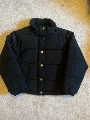 Svart pufferjacka - Snygg svart pufferjacka med hög krage och guldfärgade knappar framtill. Jackan har en boxig passform och quiltade sömmar som ger en cool look. Perfekt för kalla dagar när du vill hålla dig varm och stilren.