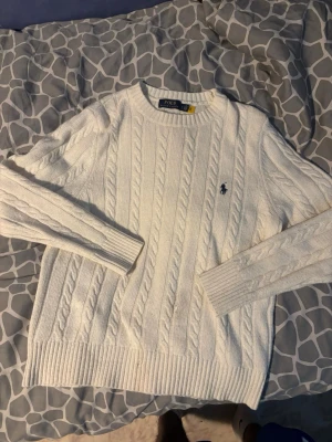 Vit kabelstickad tröja från Polo Ralph Lauren - Kabelstickad vit tröja från Polo Ralph Lauren med klassisk rund halsringning och broderad marinblå logga på bröstet. Tröjan har ribbade muddar vid ärmslut och nederkant, och är tillverkad i ett mjukt material som känns lyxigt. ‼️‼️‼️ De finns ett par fläckar på tröjan, de e på grund av fyrverkerier ‼️‼️‼️