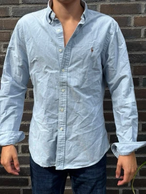 Ljusblå skjorta från Ralph Lauren - Klassisk ljusblå skjorta från Ralph Lauren i slim fit-modell. Skjortan har knappar framtill, button-down-krage och den ikoniska broderade loggan på bröstet. Tillverkad i mjuk bomull och har långa ärmar. Perfekt för en clean och stilren look.. Skriv gärna vid frågor! Modell:180/70kg