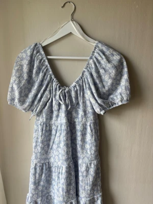Blommig blå kortklänning Gina Tricot - Supersöt blå och vit kortklänning från Gina Tricot med puffärmar och v-ringning. Klänningen har blommigt mönster, smockad resår och knyt fram. Den har bara använts en gång så har inga tecken på användning. Nypris: 399kr💕