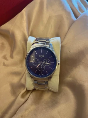 Silverfärgad klocka från Regal - Stilren klocka från Regal med blå urtavla och tre mindre subdials. Armbandet är i blankt silverfärgat metall och har ett klassiskt spänne. Perfekt accessoar för dig som gillar en clean och modern look.