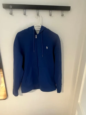 Ralph Lauren Zip Hoodie  - Blå hoodie med dragkedja och huva från Polo Ralph Lauren. Klassisk design med vit broderad logga på bröstet och snörning i huvan. Perfekt för en chill och stilren look.