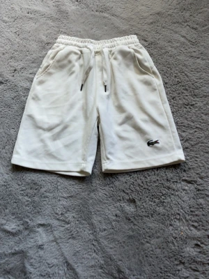 Vita shorts med snörning från Lacoste - Säljer ett par vita shorts från Lacoste med elastisk midja och snörning. Shortsen har två sidofickor och den klassiska krokodilloggan på vänster ben. Perfekta för sommaren och riktigt sköna att bära.
