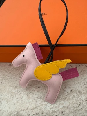 Väsksmycke rosa - Superfin Pegasus väsksmycke i ljusrosa läder med gula vingar och lila man och svans. Perfekt att hänga på väskan eller ryggsäcken för en lekfull touch. Svart läderrem för enkel fastsättning. Box ingår ej