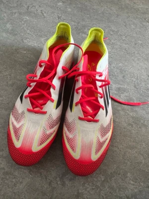 Vita och neonrosa Adidas fotbollsskor - Säljer ett par lätta fotbollsskor från Adidas i vitt med neonrosa och gula detaljer samt svarta ränder på sidorna. De är i atorlek 41,5 och aldrig använda. Finns en limegrön påse till de! 
