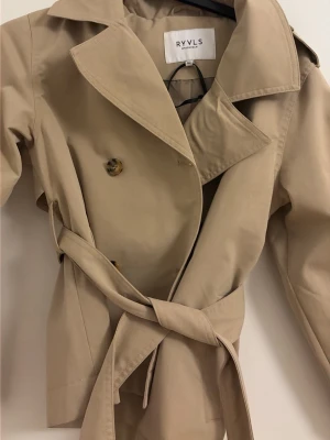 Beige trenchcoat från RYVLS(aldrig använd) - (Storlek 152 )Stilren beige trenchcoat från RYVLS med klassisk dubbelknäppning, bred krage och knytskärp i midjan. Jackan har axelklaffar, dekorativa knappar och är tillverkad i ett slätt tyg som ger en tidlös look. Perfekt för dig som vill ha en snygg och enkel trench.
