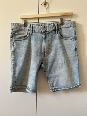 Ljusblå denimshorts från H&M i klassisk modell strl 34 - Säljer ett par ljusblå denimshorts från H&M med fem fickor och knappgylf. Shortsen har en rak passform och klassisk jeanslook med bälteshällor och slitna detaljer. Material 88% bomull 11% polyester. Storlek 34.