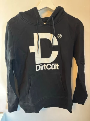 Svart hoodie från DirtCult - Svart hoodie från DirtCult med stor vit logga på bröstet och tryck på huvan. Klassisk känguruficka framtill och justerbar huva med snören. Storleken är mer som en XS/S än en M. 