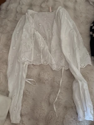 Vit broderad blus från Nelly - Söt vit blus från Nelly i storlek 34 med broderade blommor och hålmönster. Blusen har långa ärmar, knytband framtill och vågig kant nertill. Tillverkad i ett lätt och luftigt tyg, perfekt för en trendig och romantisk look.