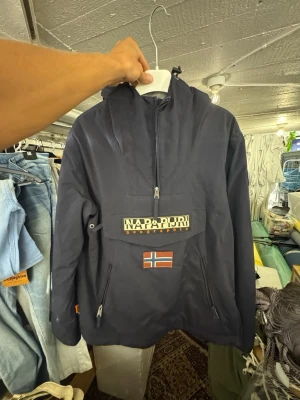 Mörkblå vindjacka från Napapijri - Snygg mörkblå vindjacka från Napapijri med stor ficka framtill, dragkedja och norsk flagga som detalj. Jackan har huva, patch på ärmen och justerbar nederkant. Perfekt för blåsiga dagar och streetwear-stil. Finns att hämta i Gröndal. Nypris är 999kr.