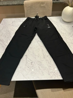 Svarta Adidas mjukisbyxor med dragsko - Svarta mjukisbyxor från Adidas med raka ben och elastisk midja med dragsko. Byxorna har två fickor med dragkedja framtill och en liten Adidas-logga på ena benet. Perfekta för en avslappnad stil.
