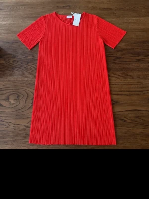 Röd plisserad kortklänning - Snygg röd kortklänning med plisserad struktur och rund hals. Klänningen har korta ärmar och rak passform, perfekt för dig som gillar en enkel men trendig look.
