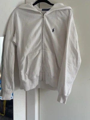 Vit hoodie från Polo Ralph Lauren - Klassisk vit hoodie från Polo Ralph Lauren med dragkedja framtill och blå logga broderad på bröstet. Hoodien har huva, kängurufickor och ribbade muddar vid ärmar och nederkant. Tillverkad i mjuk bomull för en skön och avslappnad look.