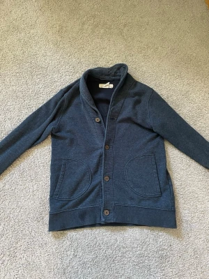 Marinblå Cardigan - Mysig mörkblå Cardigan med sjalkrage och fyra knappar framtill. Två fickor på framsidan och ribbade muddar vid ärmslut och nederkant. Perfekt för lager på lager och enkel att matcha. Storlek = 