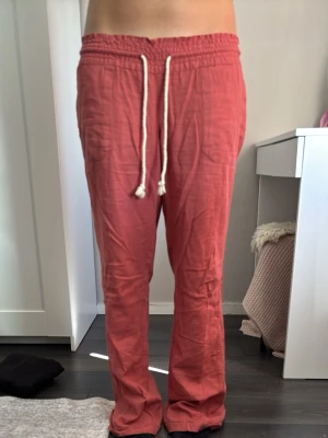 Roströda linnebyxor med dragsko - Säljer ett par roströda linnebyxor med raka ben och resår i midjan. Byxorna har vit dragsko framtill och två bakfickor. Materialet är luftigt och perfekt för varma dagar. Passar dig som gillar en avslappnad och trendig stil.