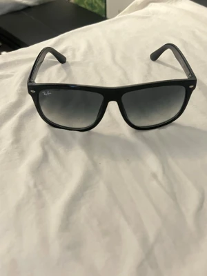 Svarta Ray-Ban solglasögon - Snygga svarta solglasögon från Ray-Ban med klassisk rektangulär form och mörka glas. Bågarna är i plast och har små silverdetaljer vid fronten. Perfekta för att höja din stil på soliga dagar.