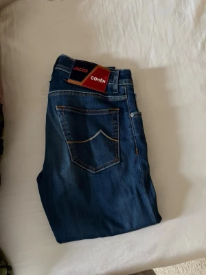 Blå jeans från Jacob Cohën - Snygga blå jeans från Jacob Cohën med klassisk femficksmodell och raka ben. Jeansen har kontrastsömmar och en patch med logga bak i midjan. Tillverkade i mjukt denimtyg som ger en skön passform.
