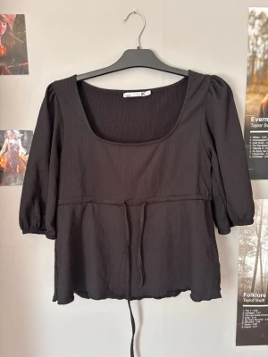 Svart blus med fyrkantig ringning - Snygg svart blus med fyrkantig ringning och trekvartsärm. Tillverkad i ett lätt, krinklat tyg och har en dekorativ knytning i midjan. Perfekt att styla med jeans eller kjol för en trendig look.