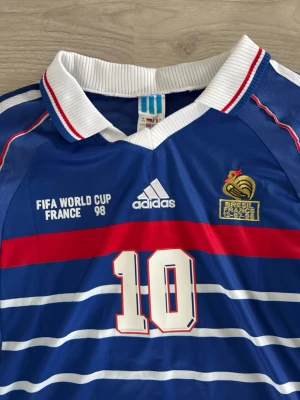 Zidane WC jersey France - Klassisk blå fotbollströja från Adidas, officiell Frankrike VM 98 med nummer 10 på bröstet. Tröjan har vita och röda detaljer, broderad tupp och FIFA World Cup-tryck. Ribbad vit krage och ränder över bröstet. Perfekt för fotbollsfans! Size M