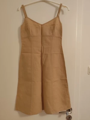 Beige ärmlös kortklänning - Snygg beige kortklänning med smala axelband och figurnära passform. Klänningen har en enkel design med sömdetaljer och dragkedja i ryggen. Perfekt för varma dagar och kan stylas på många olika sätt.
