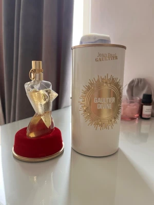 Jean Paul Gaultier Gaultier Divine parfym - Unik parfymflaska formad som en kvinnokropp i glas med gulddetaljer och guldfärgat lock. Kommer med en elegant vit och guldig förpackning där det står 'Gaultier Divine'. Flaskan står på en rund röd sammetsliknande bas.