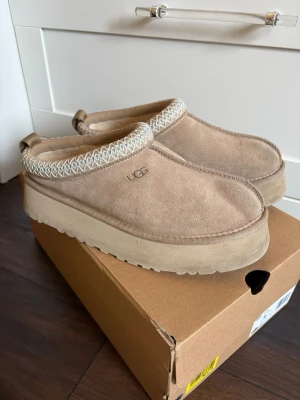 Beige UGG Tazz tofflor i mocka - Säljer ett par beige UGG Tazz tofflor i mjuk mocka med fluffigt foder och platåsula. Dekorativt mönster runt öppningen och UGG-logga på sidan. Perfekta för chill dagar hemma eller när du vill ha något extra bekvämt.