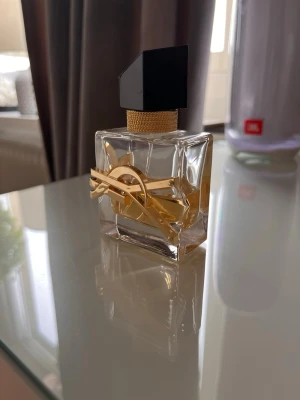 YSL Libre Eau de Parfum - Elegant parfymflaska från Yves Saint Laurent med genomskinligt glas, guldiga detaljer och svart asymmetrisk kork. Flaskan har en unik design med YSL-loggan i metall runt flaskan och innehåller en gyllene vätska.