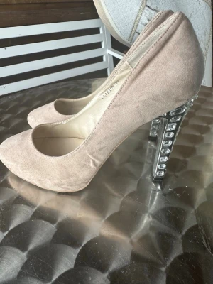 Beige/ Nude  pumps med glittrande klack - Snygga pumps i beige mockaimitation med rundad tå och hög klack dekorerad med glittrande stenar. Skorna har en klassisk siluett och ger en elegant look med extra bling tack vare klacken. Perfekta för dig som vill sticka ut lite extra.lättsmuts förekommer, klackens höjd ca 11-12 cm
