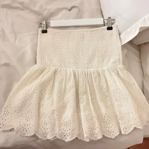 Vit kort kjol med broderade detaljer - Söt vit kortkjol med smockad midja och volangig nederdel. Kjolen har broderade spetsdetaljer och hålmönster. Perfekt för varma dagar och somriga outfits.