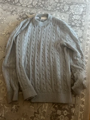 Ljusgrå kabelstickad tröja - Säljer en ljusgrå kabelstickad tröja med rund hals och ribbade muddar vid ärmslut och nederkant. Tröjan har ett klassiskt flätmönster och är perfekt för dig som gillar en stilren och mysig look. Passar till det mesta och är superenkel att matcha.