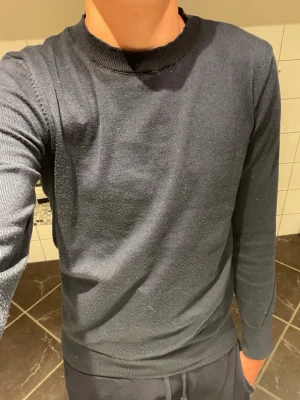 Mörkblå stickad tröja - En enkel och stilren mörkblå stickad tröja med rund halsringning och långa ärmar. Tröjan har ribbade muddar vid ärmslut och nederkant. Perfekt att matcha med jeans eller chinos för en clean look.