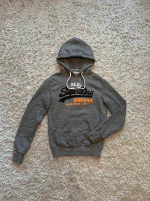Grå Superdry hoodie med tryck - Snygg grå hoodie från Superdry med svart och orange tryck på bröstet samt japanska tecken. Klassisk ficka framtill, vita dragsnören i huvan och ribbade muddar. Perfekt för en avslappnad och trendig stil.