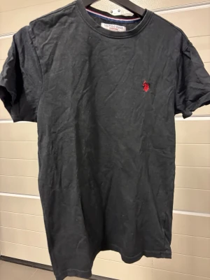 Svart t-shirt från US Polo Assn - Klassisk svart t-shirt från US Polo Assn med korta ärmar och rund hals. T-shirten har en liten röd logga broderad på bröstet och är gjord i mjuk bomull. Perfekt till jeans eller shorts för en enkel och stilren look.