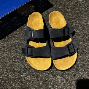 Birkenstock Arizona blå sandaler - Säljer ett par Birkenstock Arizona sandaler i mörkblått Birko-Flor material med två breda remmar och svarta spännen. Fotbädden är i naturfärgad kork och innersulan är mjuk och bekväm. Klassisk och tidlös modell som är enkel att matcha.