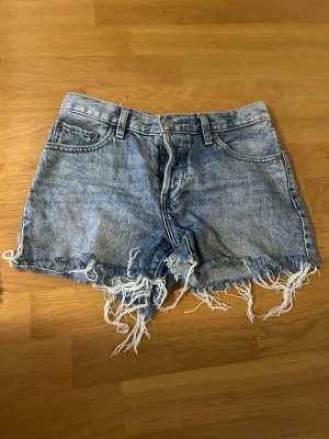 Blå jeansshorts med fransig kant - Säljer ett par klassiska blå jeansshorts med rå, fransig kant och femficksmodell. Shortsen har hög midja och är tillverkade i slitstark bomull. Perfekta för sommardagar och ger en avslappnad vibe. Har en fläck på insidan av ägaren innan mig, fråga om bild. Strl 32 funkar som 34