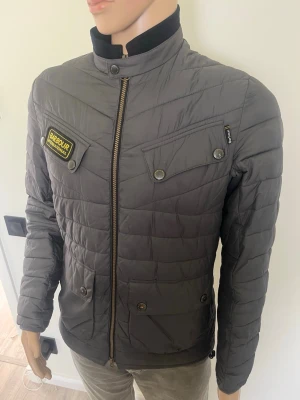 Grå Barbour pufferjacka storlek M - Snygg grå pufferjacka från Barbour med quiltat mönster, hög krage och dragkedja framtill. Jackan har två bröstfickor med knappar, två sidofickor och en Barbour International-patch på bröstet. Perfekt för kalla dagar och riktigt stilren look.