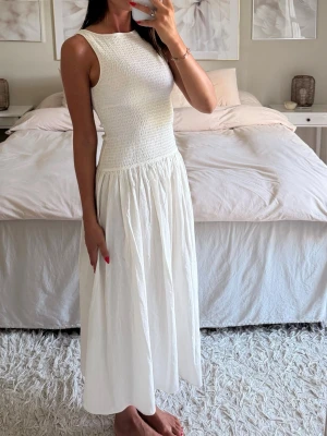 Smockad cremefärgad klänning H&M  - Elegant vit långklänning med smockat liv och rund halsringning. Klänningen har ärmlös design och ett luftigt, lätt utställt kjolparti. Perfekt för varma dagar och somriga tillfällen. Tillverkad i ett mjukt, tunt material som ger en skön känsla.