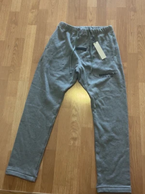 Grå Essentials mjukisbyxor - Snygga grå mjukisbyxor från Essentials Fear of God med dragsko i midjan och tryckt logga på vänster lår. Byxorna har raka ben och elastisk midja för skön passform. Perfekta för chill dagar och streetwear-stil.